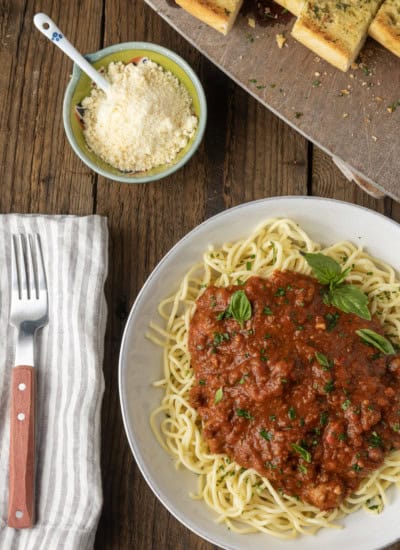 spaghetti sauce on spagetti on a plate topped by fresh herbs with a napkin and fork on the left and garlic bread on a cutting board above.