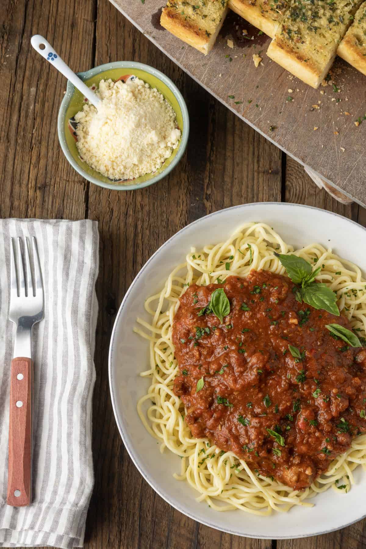 spaghetti sauce on spagetti on a plate topped by fresh herbs with a napkin and fork on the left and garlic bread on a cutting board above.