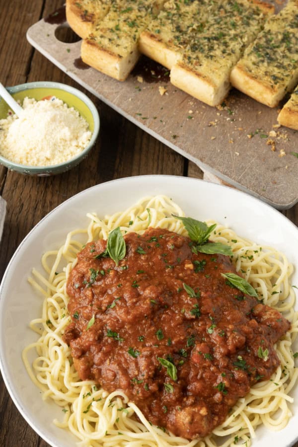 spaghetti sauce on spagetti on a plate topped by fresh herbs with a napkin and fork on the left and garlic bread on a cutting board above.