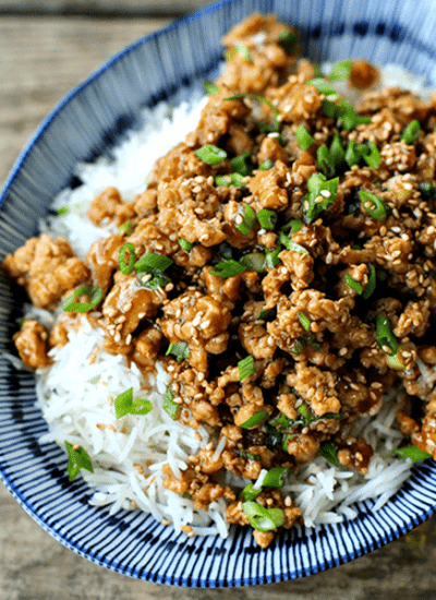 Cheater Sesame Chicken ground chicken in sweet and spicy sauce, sesame seeds, sliced green onions, long grain white rice, asian bamboo pattern blue and white bowl on wooden bench